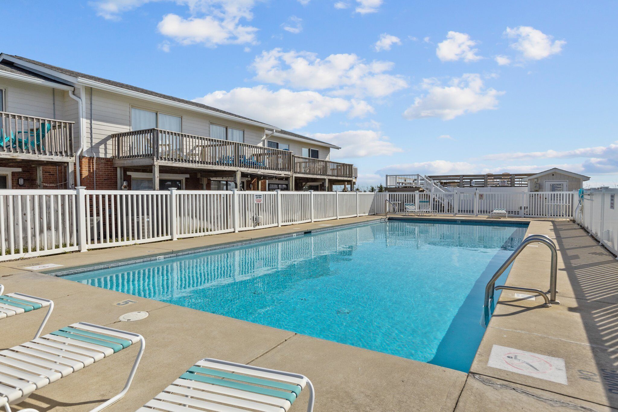 A swimming pool with lounge chairs, a white fence, and a building with balconies.