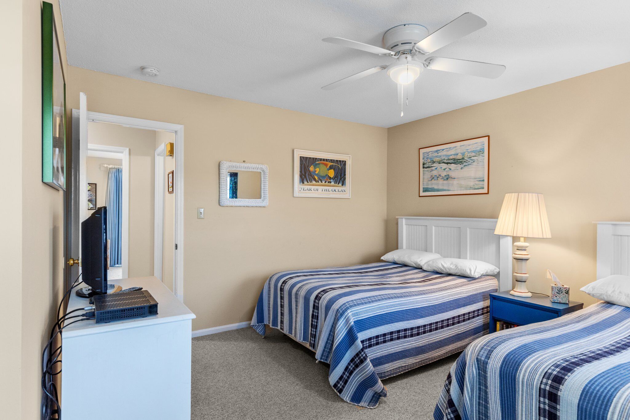 A bedroom with two beds, a ceiling fan, a desk with a computer, and artwork on the walls.