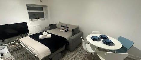 A compact room with a bed, a sofa, a dining table with chairs, a television, and a window.
