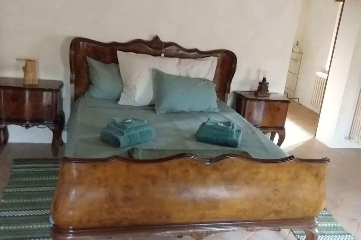 Een bed met houten frame en een groene spreidek, twee kussens en opgevouwen handdoeken op een kamerkleed met dessin.