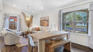 A modern living room with a wooden dining table, white sofas, and a large window with a view of the street. - Pool, 3-min to Main, Sleeps 8 (Fredericksburg)
