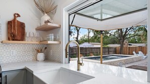 A modern kitchen with a large sink, wooden shelves, and a view of a backyard with a pool and outdoor seating. - Pool, 3-min to Main, Sleeps 8 (Fredericksburg)