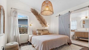 A bedroom with a large bed, a hanging light fixture, a window with curtains, a wooden floor, and a bathroom with a mirror and sink. - Pool, 3-min to Main, Sleeps 8 (Fredericksburg)