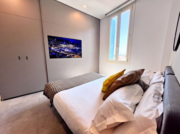 Un dormitorio con cama, almohadas, una mesita de noche, una ventana y un televisor empotrado que muestra una vista urbana.