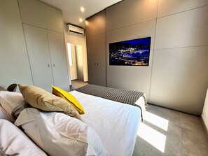 Un dormitorio moderno con una cama, un ventanal grande, un equipo de aire acondicionado y un cuadro con un paisaje urbano en la pared.