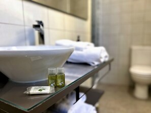 Un baño con un lavamanos grande de color blanco, dos botellas de aceite de oliva y una pastilla de jabón sobre una repisa de vidrio.