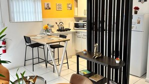 Een moderne woonkamer met een bank, een eethoek met tafel en stoelen, een keuken met apparatuur, en een decoratieve zwarte scheidingswand.