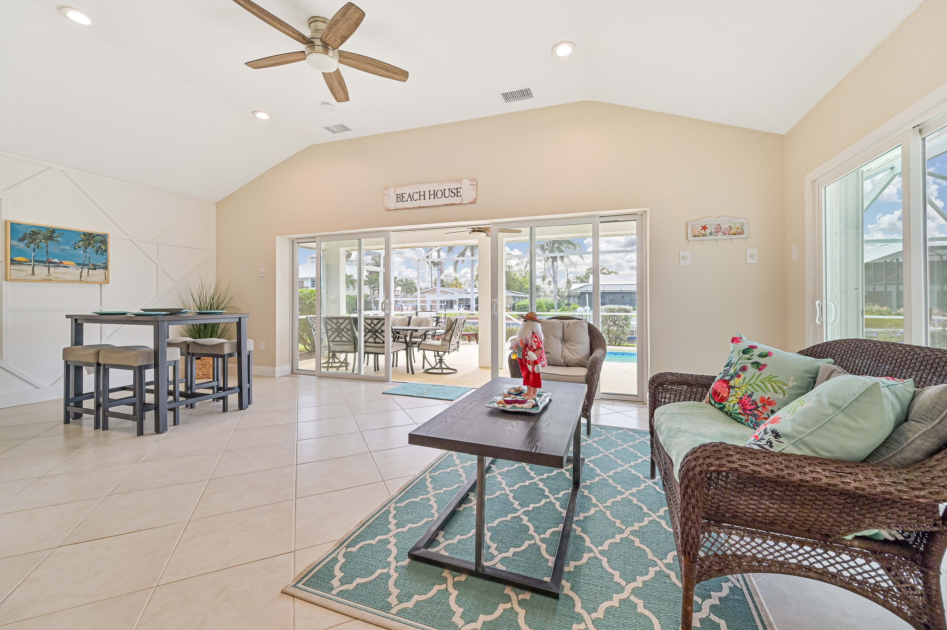 A spacious living area with a ceiling fan, a dining table with chairs, a wicker sofa with cushions, and a large window offering a view of the outdoors.