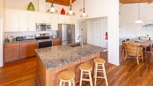 A kitchen with granite countertops, wooden cabinets, stainless steel appliances, and a central island with stools. - Brownley Beach Cottage- steps to the Beach, Elevator, Dog Friendly! (Virginia Beach)