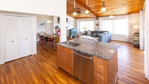 A spacious kitchen with wooden cabinets and a granite countertop, a dining area with a table and chairs, and a living room with a sofa and a TV. - Brownley Beach Cottage- steps to the Beach, Elevator, Dog Friendly! (Virginia Beach)