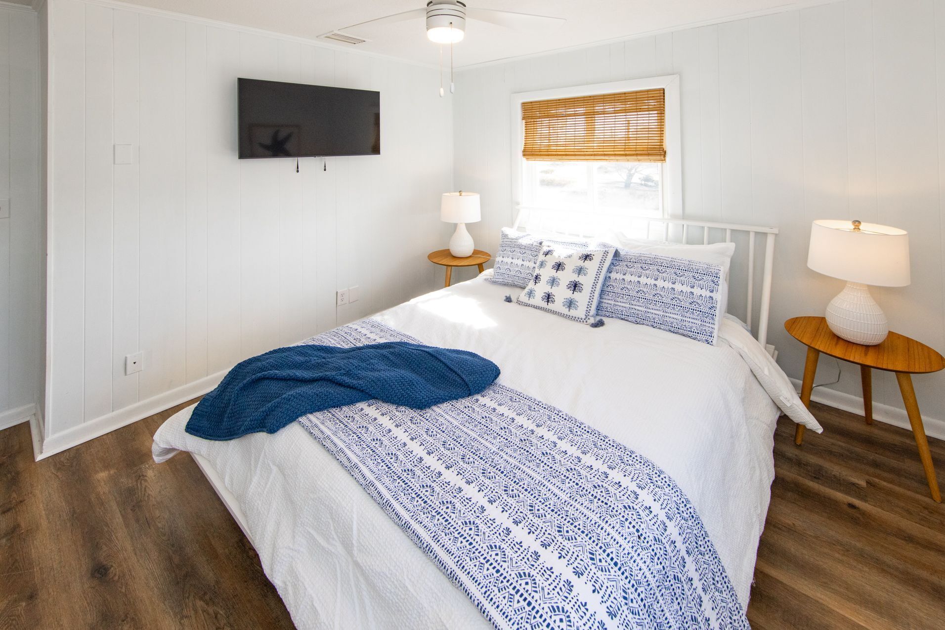 A bedroom with a bed, two bedside tables, a TV mounted on the wall, a window with blinds, and a ceiling fan.