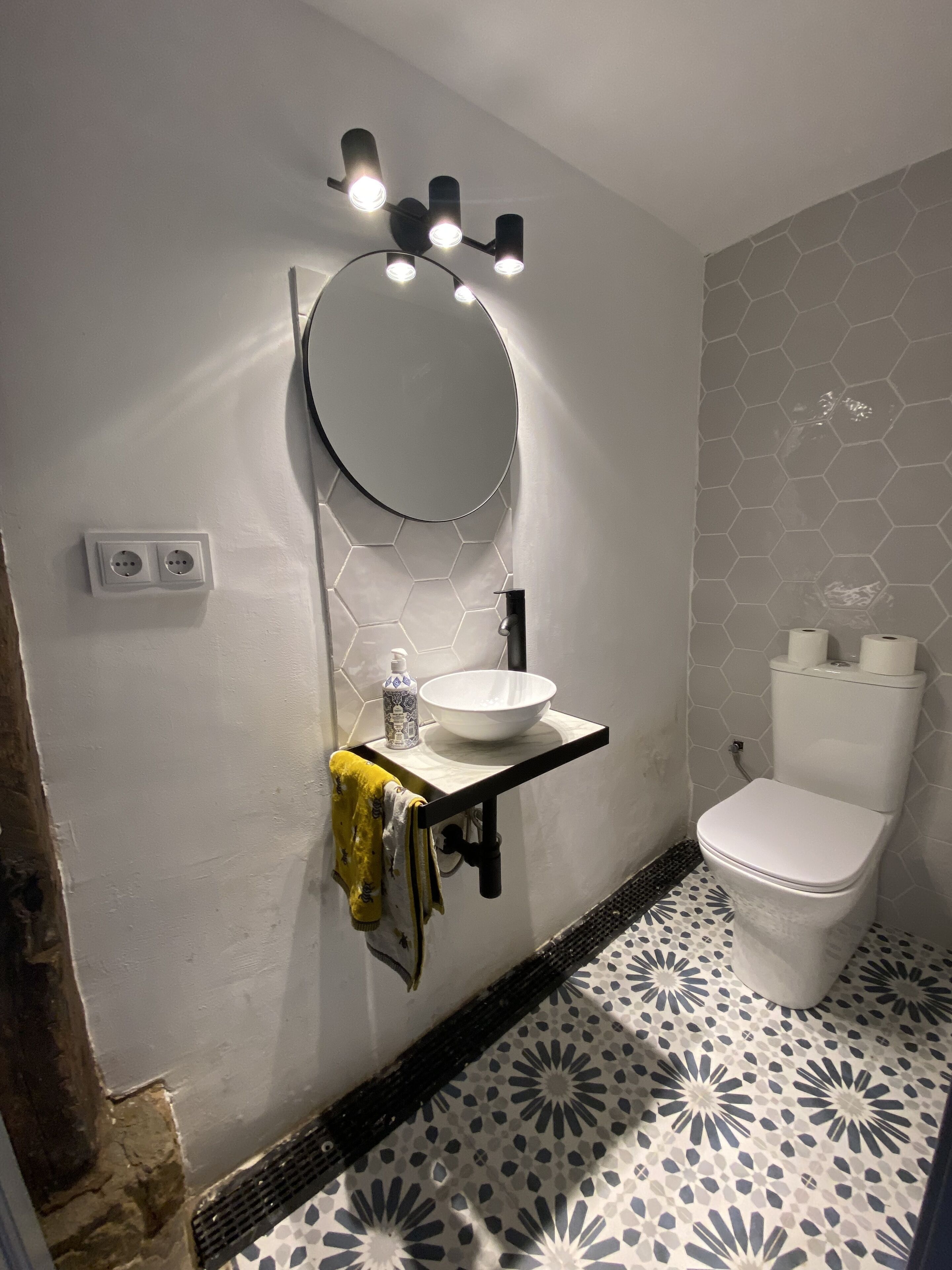 A bathroom with a white sink, a mirror, and a toilet. There are two electrical outlets on the wall, and a towel is hanging on the side.
