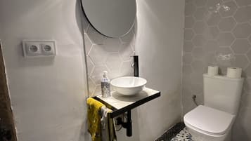 A bathroom with a white sink, a mirror, and a toilet. There are two electrical outlets on the wall, and a towel is hanging on the side.