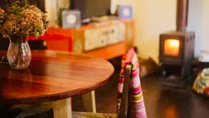 A cozy room with a wooden table, a chair with a colorful cushion, a vase with dried flowers, a television, and a wood-burning stove. - Escalera al puerto Guest House  (Valparaiso)