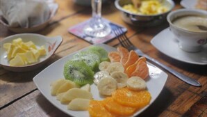 A plate of sliced fruits including kiwi, banana, and orange, with a fork and a glass of orange juice on a wooden table. - Escalera al puerto Guest House  (Valparaiso)