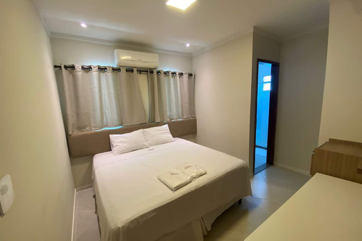 Quarto com cama, ar condicionado, janela com cortinas e porta que dá para outro cômodo.