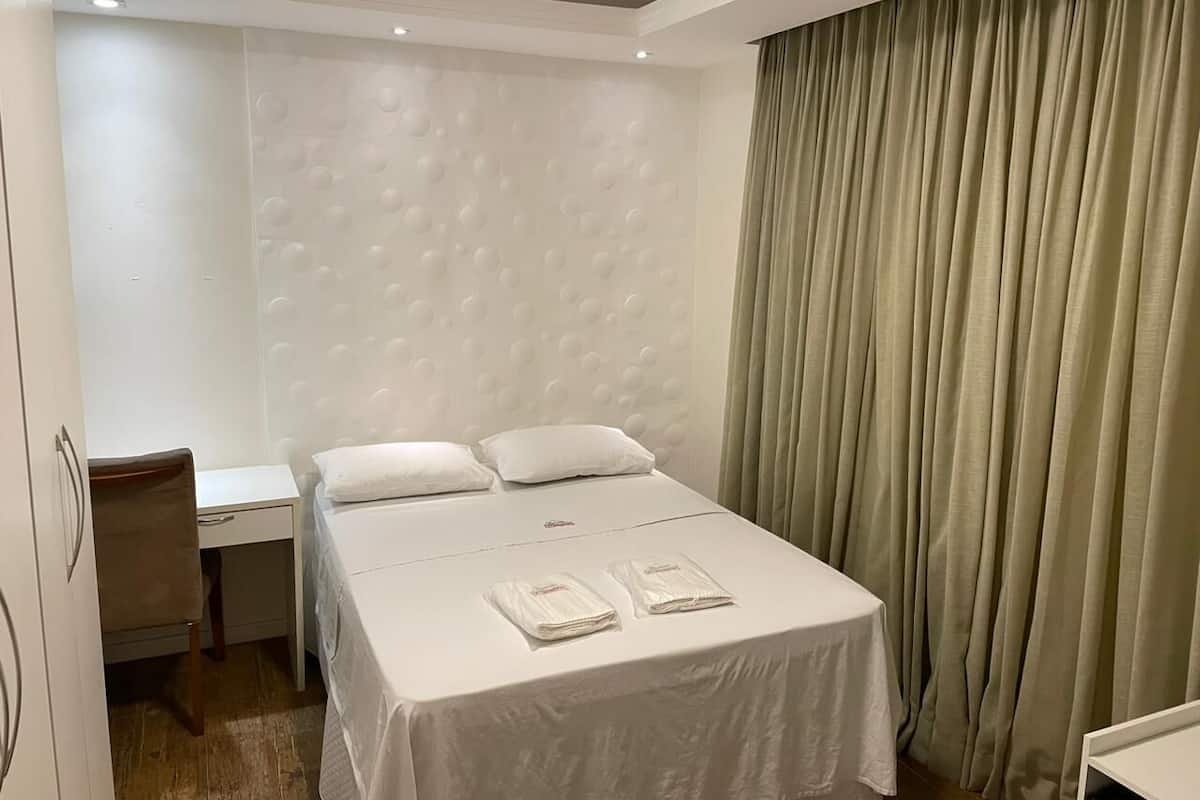 Quarto de hotel com uma cama, uma escrivaninha e uma cadeira. Há duas toalhas sobre a cama.