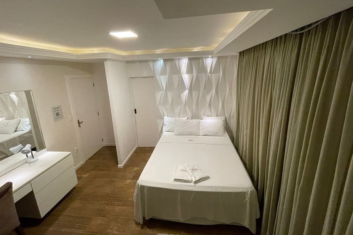 Quarto de hotel com cama grande, mesa de cabeceira, espelho e cortina.