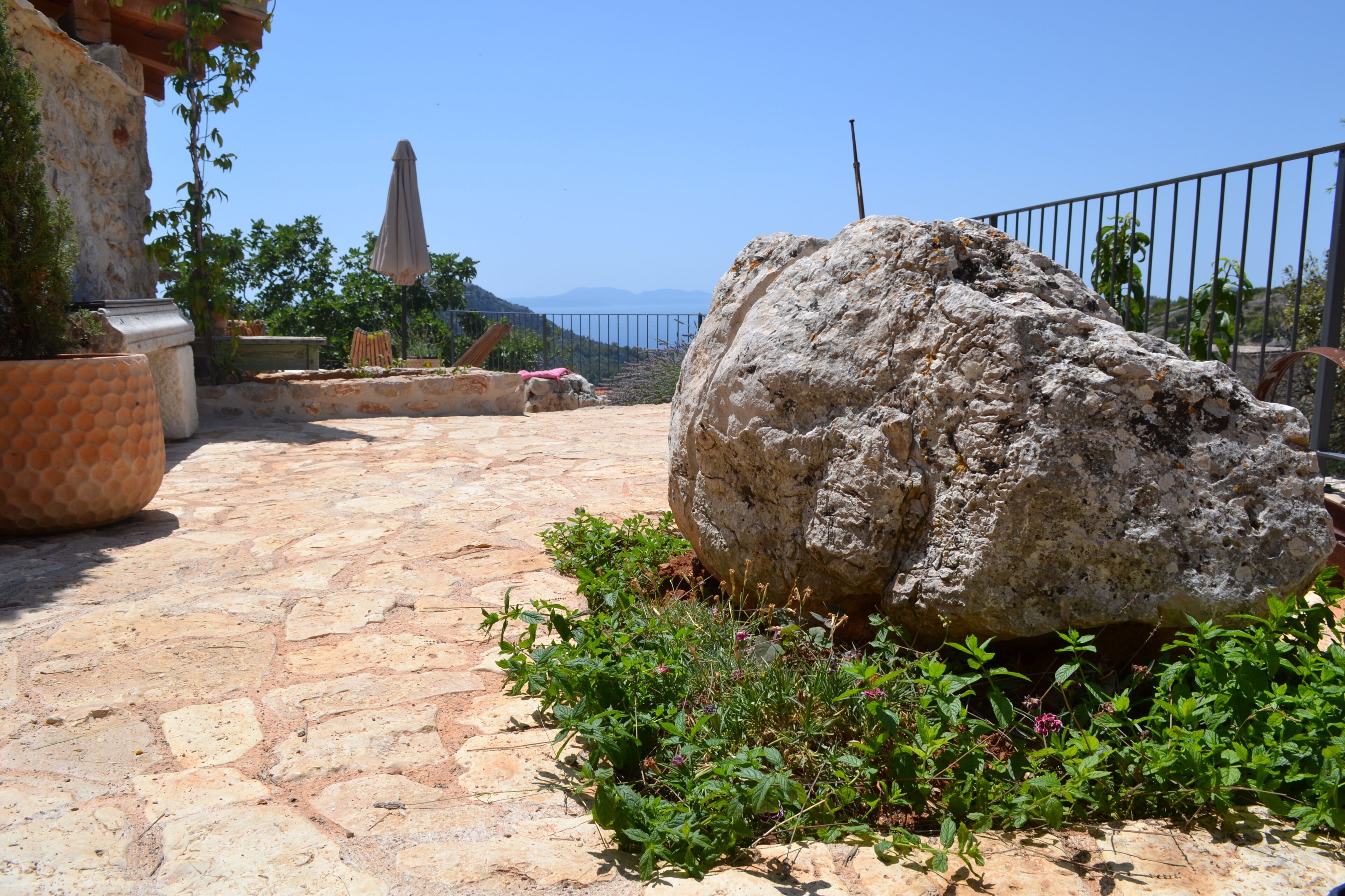 A stone-paved patio with a large rock, potted plants, and a metal fence.