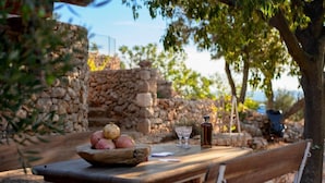 A wooden table with a bowl of fruit, two glasses, and a bottle, set outdoors with stone walls and trees in the background. - Secluded and Peaceful Retreat House on the Island of Hvar (Velo Grablje)