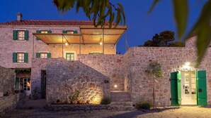 A stone building with a red-tiled roof, green shutters, and a well-lit entrance. - Secluded and Peaceful Retreat House on the Island of Hvar (Velo Grablje)