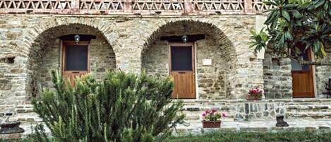 A stone building with arched doorways and windows, a balcony with a lattice design, and potted plants.