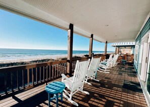 Una terraza techada con piso de madera, sillas mecedoras blancas y una pequeña mesa, con vista a la playa.