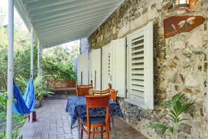 A covered outdoor seating area with a stone wall, wooden chairs, and a table with a patterned tablecloth.