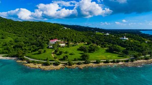 Vue aérienne d’une propriété côtière avec une grande maison entourée d’une végétation luxuriante et sous un ciel bleu dégagé.