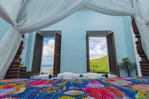Un dormitorio con un edredón colorido, una cama con toallas blancas y vistas al mar a través de contraventanas abiertas.