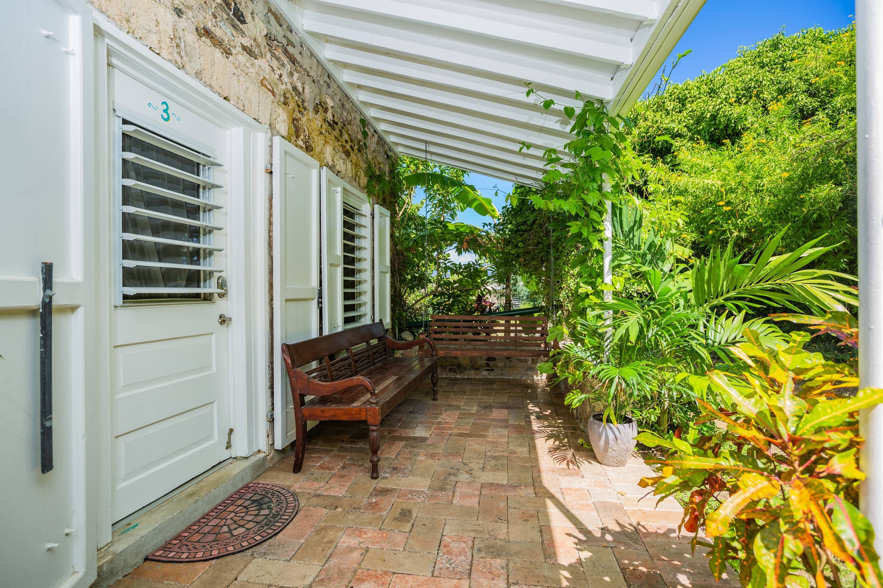 Une terrasse couverte avec un banc, une porte à volets et des plantes en pot.