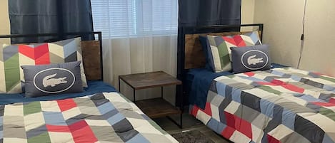 Une chambre avec un grand lit et deux lits simples, chacun recouvert d’une courtepointe aux couleurs vives, ainsi qu’une petite table et une lampe.