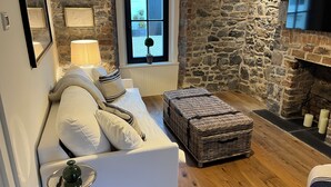 A cozy room with a stone wall, a white sofa with cushions, a wicker ottoman, a lamp, and a fireplace. - In Town Beautiful Trim Castle Cottage   (Trim)