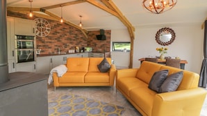 A modern kitchen with a brick wall, wooden beams, and a large clock. There are two yellow sofas with cushions, a patterned rug, and a dining area with a table and chairs.