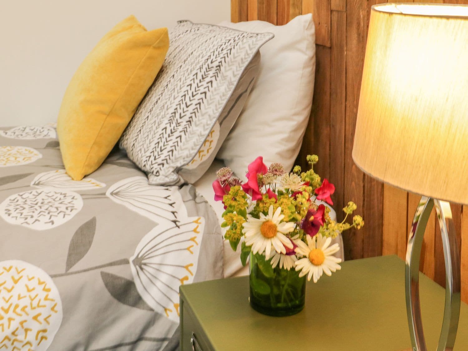Una cama bien tendida con ropa de cama estampada, una almohada amarilla, una almohada decorativa, una mesita de noche verde con una lámpara y un florero con flores.