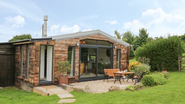 A small wooden cabin with a glass door and windows, a patio with a table and chairs, and a garden with plants and a pathway.