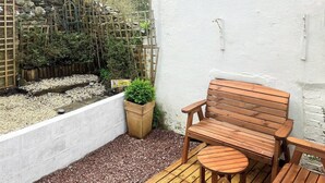 Una terraza de madera con un banco y una mesa, rodeada por un muro blanco y un jardín con plantas y un letrero de advertencia.