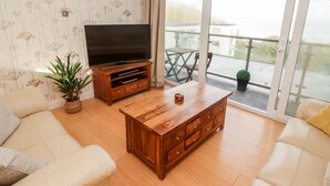 A living room with a wooden coffee table, a sofa, a TV on a wooden stand, and a balcony with a view. - 54 Croft Court (Tenby)