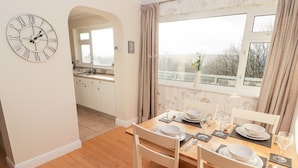 A dining area with a wooden table set for four, white chairs, and a large wall clock. - 54 Croft Court (Tenby)