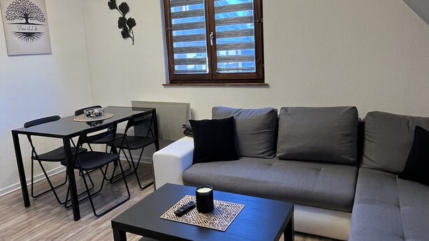 A modern living room with a sectional sofa, a dining table with chairs, a coffee table with a candle, and a ceiling light fixture. - Appartement F2 Duplex Turckheim, Alsace Situé sur la Route des Vins (Turckheim)