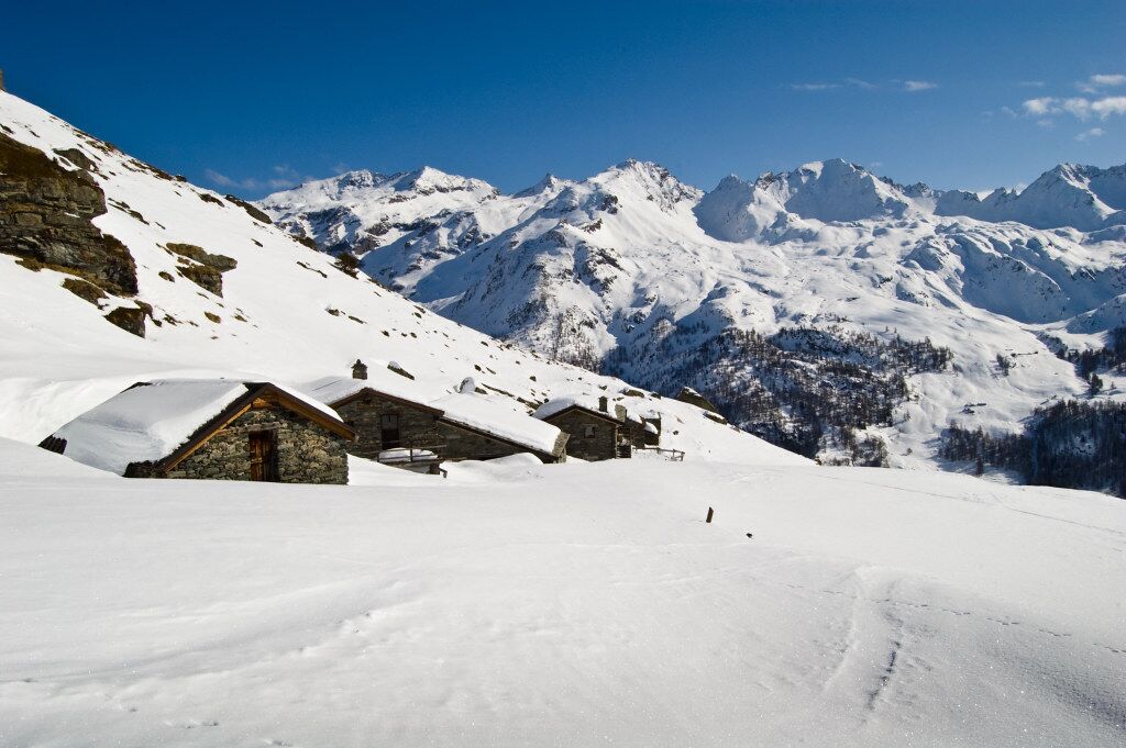Ein verschneites Bergdorf mit Steinbauten und freier Sicht auf ferne Berggipfel.