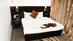 Una cama bien hecha con un almohadón marrón, uno blanco y un adorno encima. Junto a la cama hay una mesita de noche con un teléfono y una lámpara. La habitación tiene un cabecero de madera y una cortina en la ventana.