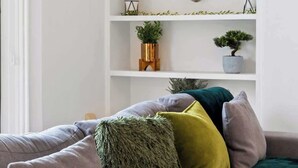 Ein Wohnzimmer mit grauem Sofa, mehreren Kissen, einem weißen Regal mit Dekoration und einer Ziegelwand.