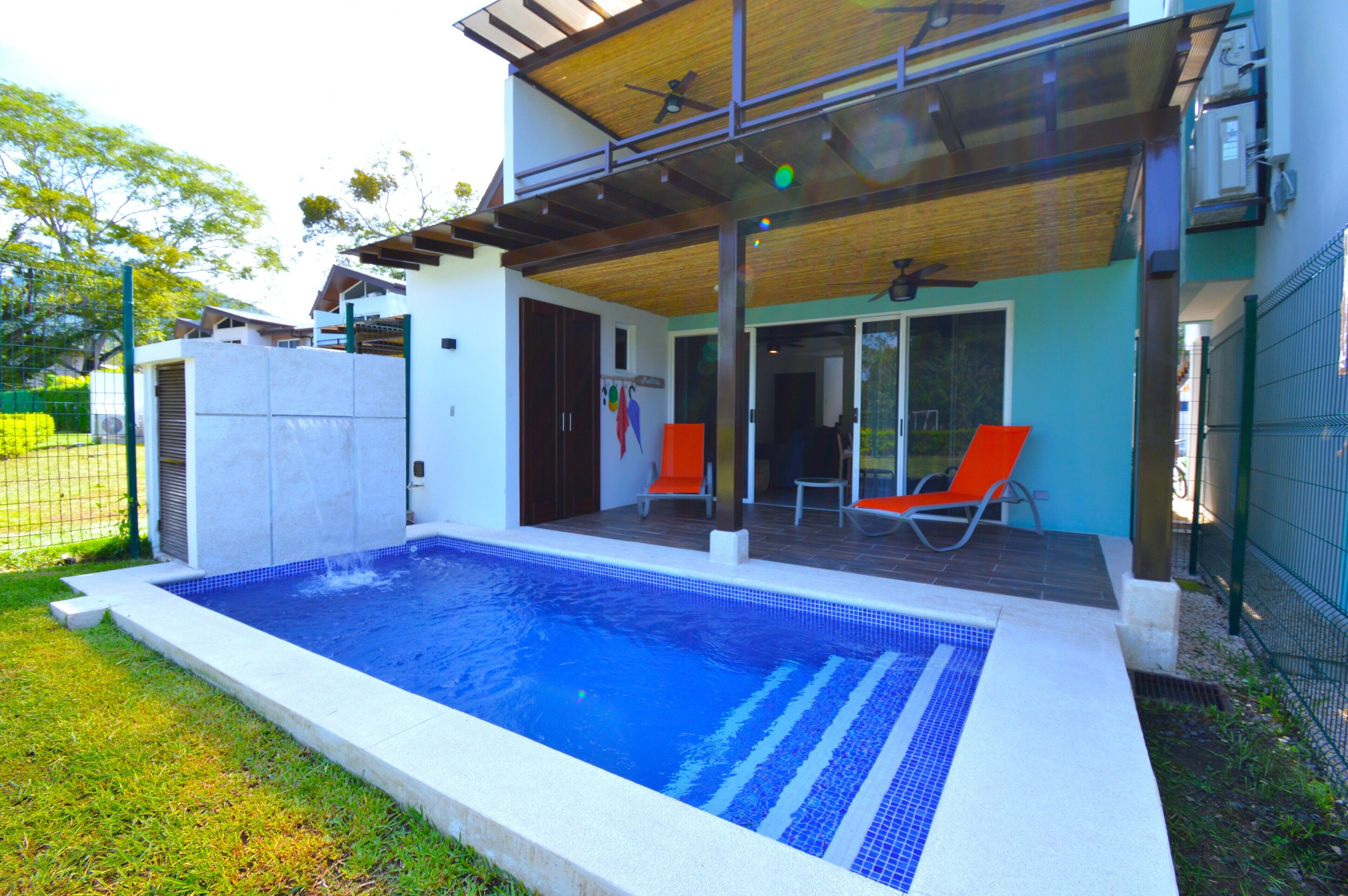 Piscina com área de estar coberta e varanda adjacente a um edifício com porta de correr de vidro.