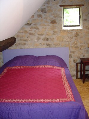 Quarto com parede de pedra, janela, cama com colcha roxa e vermelha e cabeceira de madeira.