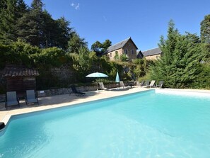 Piscina com espreguiçadeiras e guarda-sóis, cercada por árvores e um edifício de pedra ao fundo.