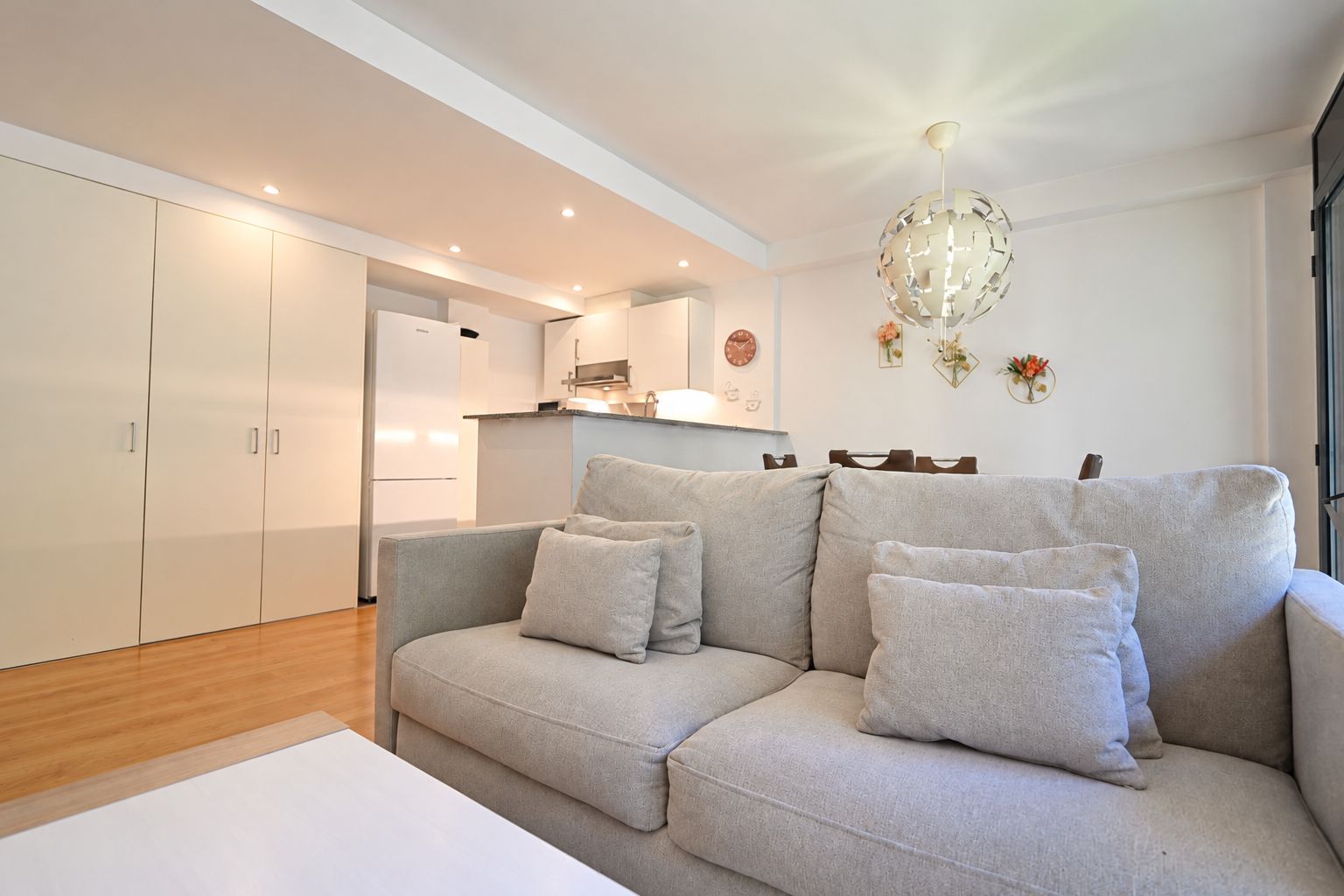 Een moderne woonkamer met een grijze bank, witte muren en een keuken op de achtergrond.