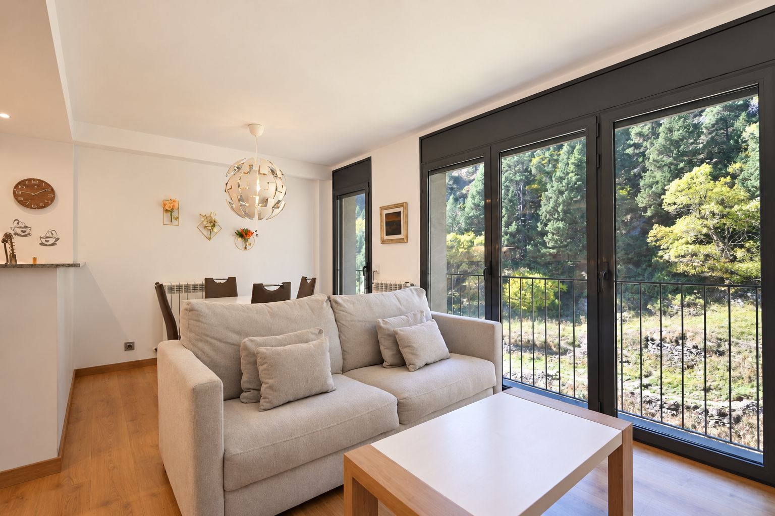 A modern living room with a sofa, a coffee table, and large windows offering a view of a forested area.