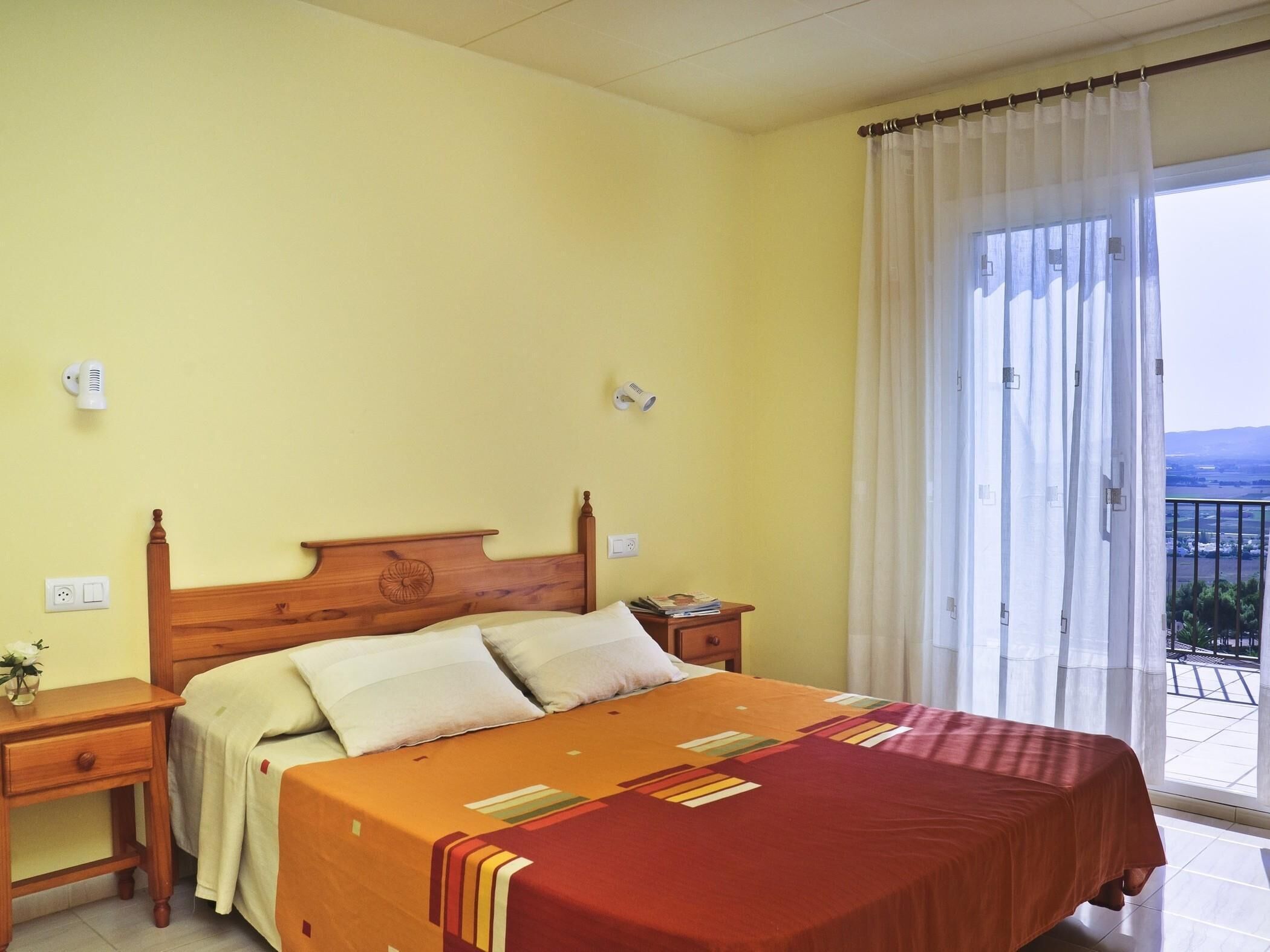 Une chambre à coucher avec un lit en bois, une table de chevet, une commode et une vue sur l’extérieur grâce à une grande fenêtre agrémentée de rideaux.