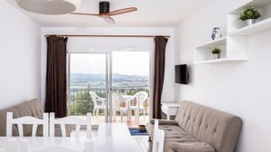 A modern living room with a dining area, a sofa, a TV mounted on the wall, and a balcony with a view of the countryside. - APCOSTAS Club Torre Vella / One Bedroom (L'Estartit)